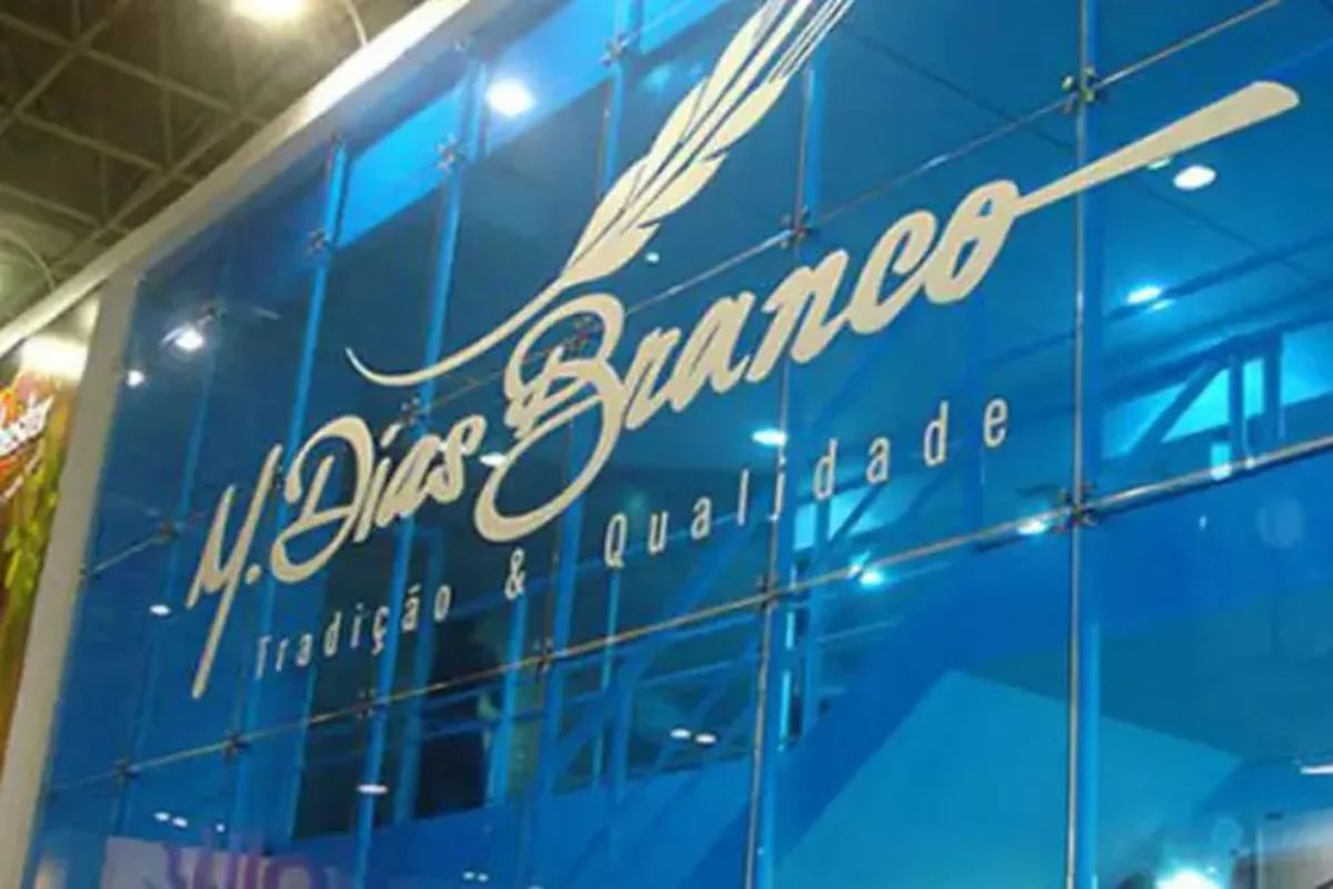 Logotipo da empresa M. Dias Branco em destaque em uma superfície de vidro azul. A logomarca inclui o nome "M. Dias Branco" em letras cursivas brancas, com uma pena estilizada acima, e o slogan "Tradição & Qualidade" em letras menores.