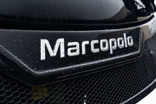 Logo do carro Marcopolo em uma grade dianteira com gotas d'água, em close-up, realçando detalhes e textura.