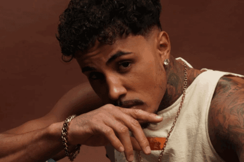 Retrato em plano médio de MC Livinho, com cabelo cacheado, barba rala e diversas tatuagens no pescoço e braços. Ele usa uma regata branca e uma pulseira dourada grossa, olhando diretamente para a câmera