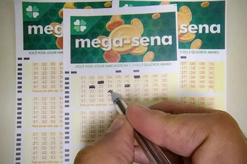 Uma foto em close-up mostra uma mão segurando uma caneta, marcando números em um bilhete de loteria da Mega-Sena (uma loteria brasileira).