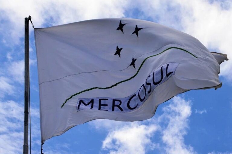 Bandeira oficial do Mercosul, de cor branca, tremulando contra um céu azul com nuvens. O design apresenta quatro estrelas pretas em semicírculo sobre duas linhas curvas (uma cinza e uma verde) e o nome 'MERCOSUL' em letras maiúsculas escuras na parte inferior.
