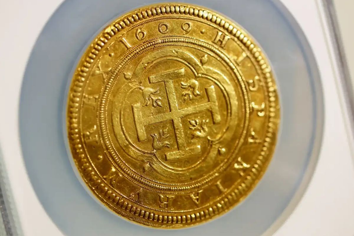 Close-up de uma moeda de ouro antiga, exibindo uma cruz gravada no centro e o ano '1609' e inscrições visíveis ao redor da borda, protegida por um suporte de plástico.