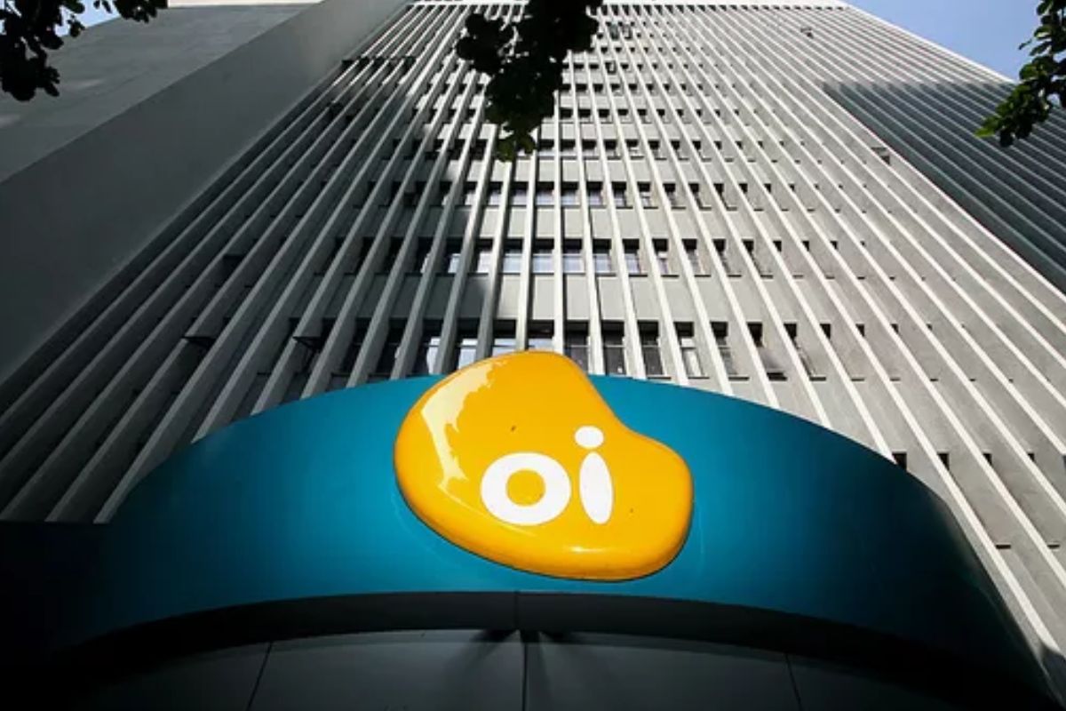 Uma vista em ângulo baixo da fachada de um prédio alto e moderno, com muitas janelas verticais. Na base do prédio, em uma marquise azul, está o grande logotipo da Oi – uma forma orgânica amarela brilhante com as letras 'oi' em branco.
