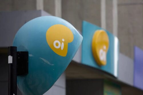 Orelhão (telefone público) e letreiro da Oi (OIBR3) na cor azul-claro e amarelo. O orelhão está em primeiro plano, inclinado, com o logo "oi" em branco dentro de uma forma amarela. O letreiro está desfocado ao fundo, também com o logo da Oi.