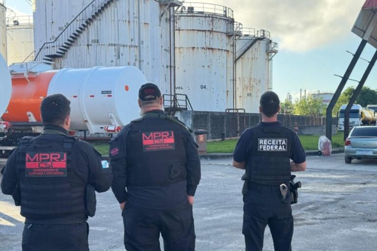 Agentes do MPRJ (Ministério Público do Rio de Janeiro) e da Receita Federal em coletes pretos, de costas, inspecionando tanques de armazenamento de combustível.