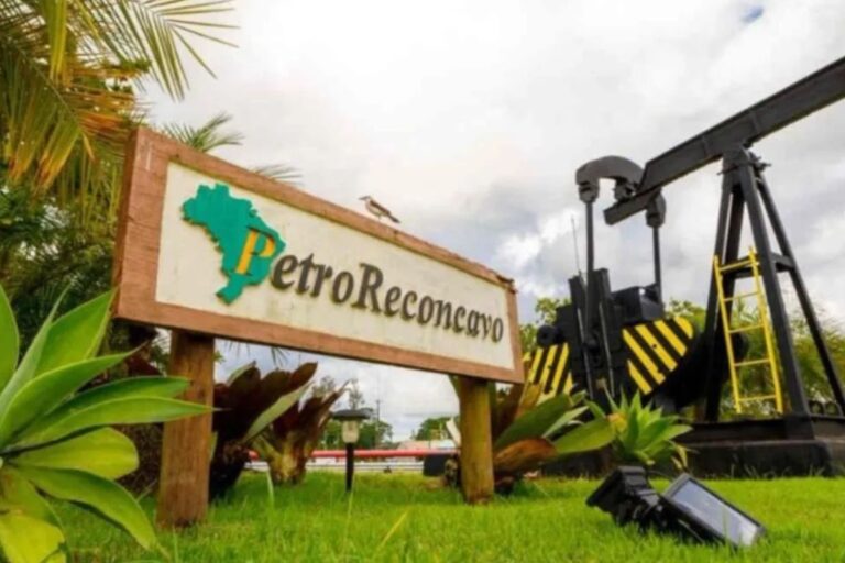 Placa de entrada da PetroReconcavo em uma área verde. A placa de madeira rústica exibe o nome 'PetroReconcavo' e o mapa do Brasil estilizado em verde, representando o Nordeste.