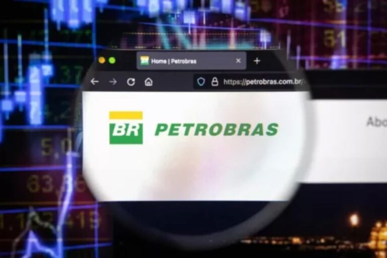 A página inicial do site oficial da Petrobras, exibindo seu logotipo verde e amarelo em um navegador web, é vista através de uma lupa ou lente circular. O fundo da imagem está desfocado, mas visivelmente mostra gráficos de mercado de ações e dados financeiros em tons de azul e roxo