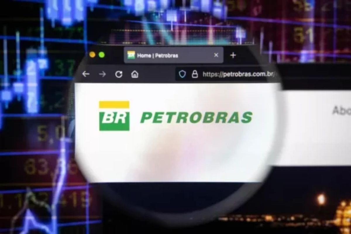A página inicial do site oficial da Petrobras, exibindo seu logotipo verde e amarelo em um navegador web, é vista através de uma lupa ou lente circular. O fundo da imagem está desfocado, mas visivelmente mostra gráficos de mercado de ações e dados financeiros em tons de azul e roxo