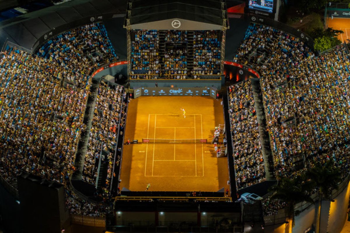Vista aérea noturna da quadra central de tênis de saibro (terra batida) no Rio Open, com um jogador no centro e as arquibancadas lotadas de público sob luzes fortes e artificiais.