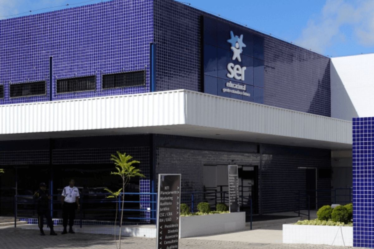 Fachada moderna do edifício da Ser Educacional. A parte superior é revestida com azulejos azuis e exibe o logotipo e o nome da empresa em branco. A entrada principal, com painéis escuros e um toldo branco horizontal, tem dois indivíduos uniformizados parados à esquerda e um jardim lateral.