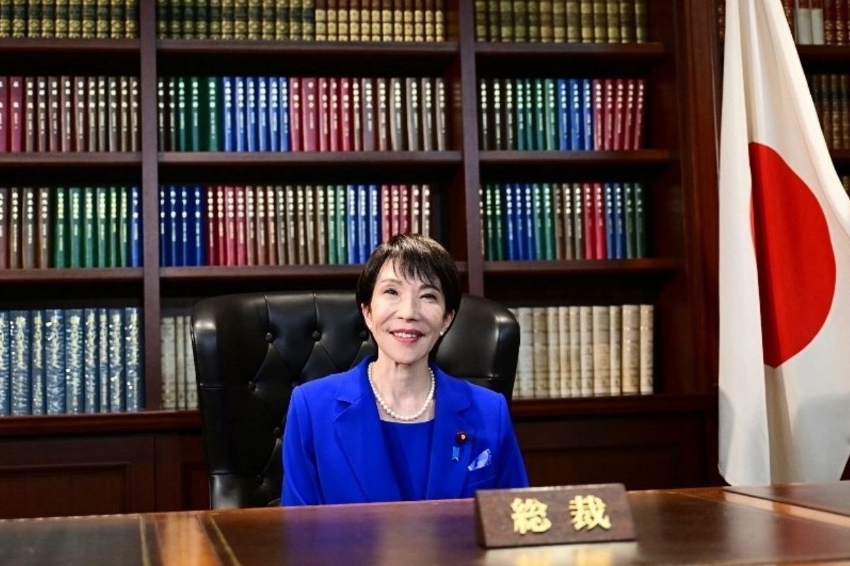 Retrato da política japonesa Sanae Takaichi, sorrindo e vestindo um tailleur azul-royal e um colar de pérolas, sentada em uma grande cadeira de couro atrás de uma mesa de madeira.