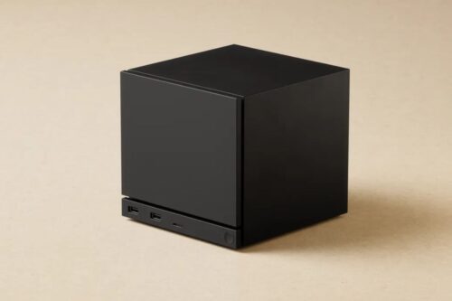 Uma Steam Machine com formato cúbico e cor preta fosca, colocado sobre uma superfície bege neutra. O dispositivo é compacto e minimalista. Na parte inferior frontal, é possível ver duas portas USB, uma pequena luz indicadora e o botão de energia.