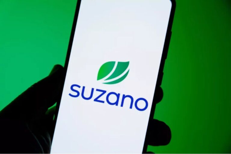 O logotipo da empresa Suzano, que consiste no nome 'suzano' em letras minúsculas azuis e um símbolo de duas folhas verdes, é exibido em uma tela de smartphone. Uma mão segura o telefone, e o fundo é um verde vibrante e escuro.
