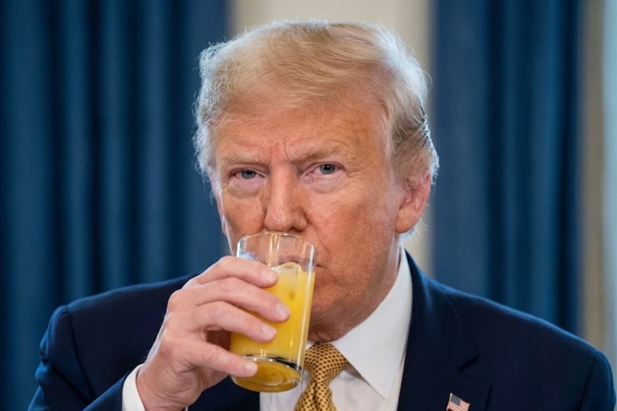 Donald Trump vestindo terno azul escuro e gravata amarela, segurando um copo de vidro com suco de laranja próximo à boca.
