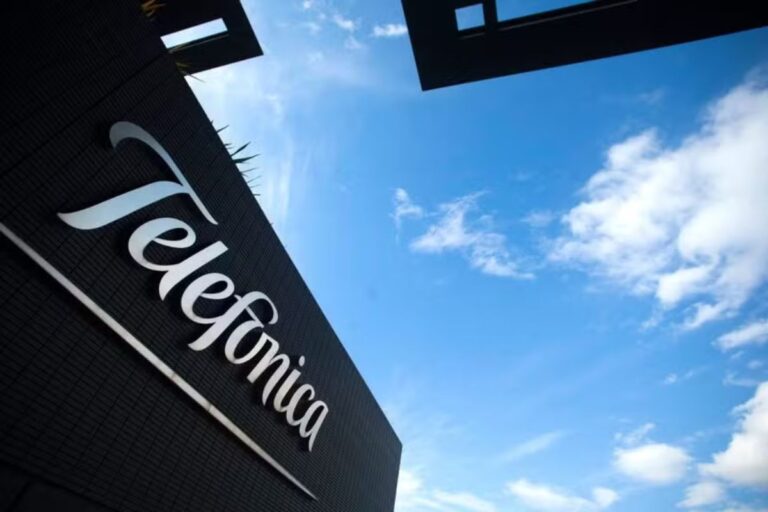 Perspectiva de baixo para cima mostrando o logotipo curvo e estilizado "Telefonica" em branco, fixado na fachada escura e moderna de um edifício corporativo. O céu está azul com algumas nuvens brancas, e a luz sugere um dia claro.