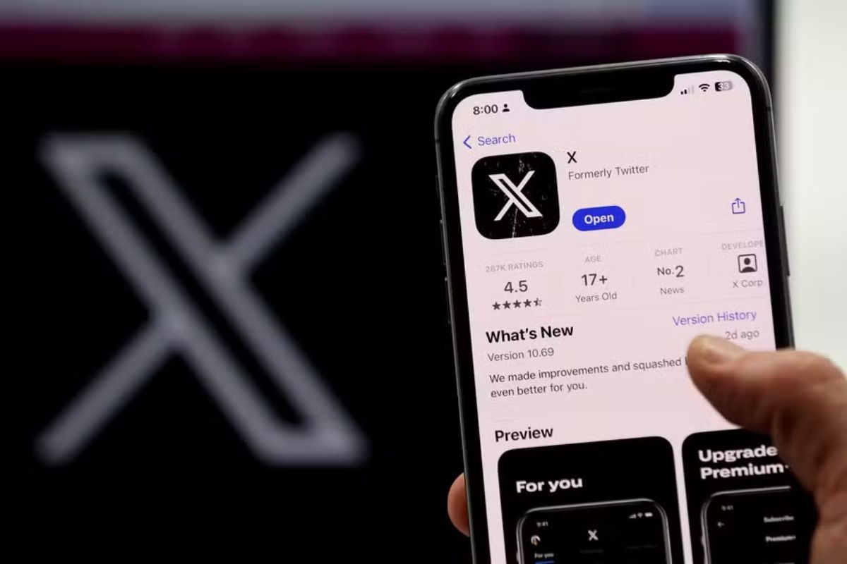 Página do aplicativo 'X, Formerly Twitter' (Antigo Twitter) exibida na App Store em um smartphone segurado. A tela mostra o ícone do novo logotipo da letra 'X'. Ao fundo, vê-se uma projeção grande e desfocada do mesmo logotipo 'X' em branco sobre um fundo escuro.