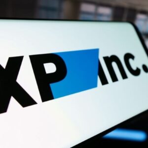 Close-up da tela de um dispositivo móvel exibindo o logotipo da empresa XP Inc. O logo é composto pelas letras 'XP' e 'inc.' na cor preta, separadas por uma forma geométrica azul. O fundo da imagem mostra um ambiente de escritório moderno e desfocado.