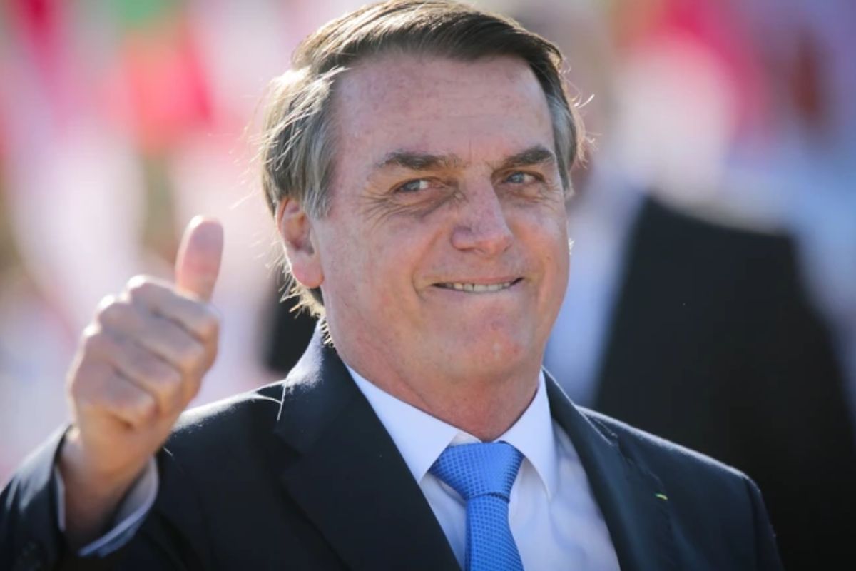 Close-up do político brasileiro Jair Bolsonaro, vestindo um terno escuro e gravata azul, sorrindo e fazendo um sinal de positivo com a mão direita levantada.