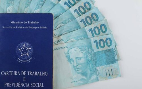 cédulas de 100 reais e uma carteira de trabalho, ilustrando a matéria sobre o décimo terceiro salário