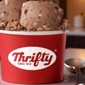 Sorvete Thrifty Ice Cream em pote vermelho com logo da marca e colher ao lado em ambiente iluminado.