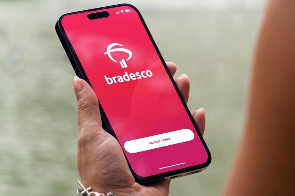 Pessoa segurando um smartphone com o aplicativo do Bradesco aberto na tela inicial. O fundo do app é vermelho-rosado com a logo do Bradesco em branco e um botão escrito "acessar conta" na parte inferior.