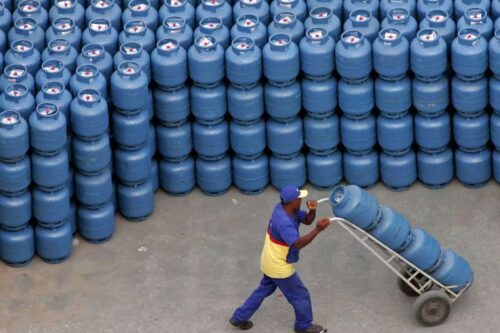 A imagem mostra uma vista aérea de um grande pátio de distribuição com centenas de botijões de gás de cozinha azuis empilhados em fileiras.