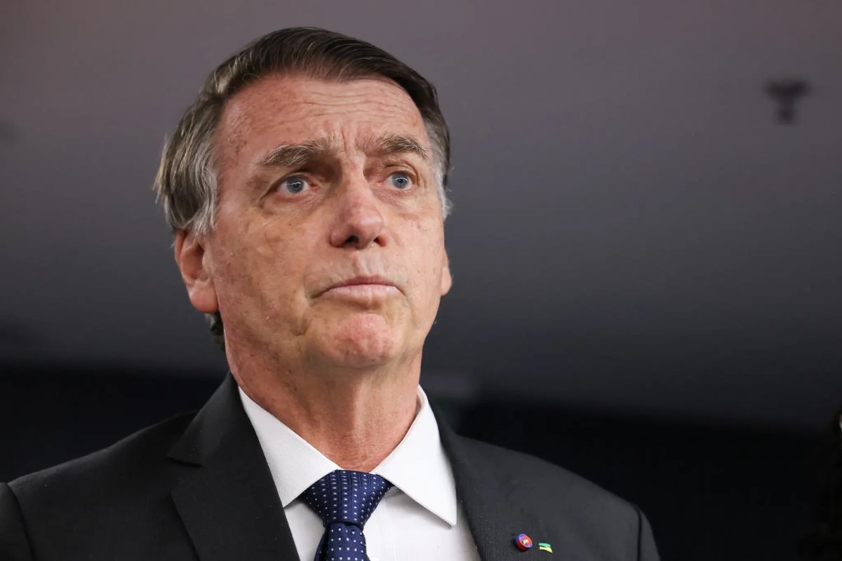 Fotografia em close do ex-presidente Jair Bolsonaro. Ele veste um paletó escuro, camisa branca e gravata azul com pequenos detalhes. Bolsonaro está com uma expressão séria, olhando levemente para cima e para o lado, contra um fundo neutro e desfocado.