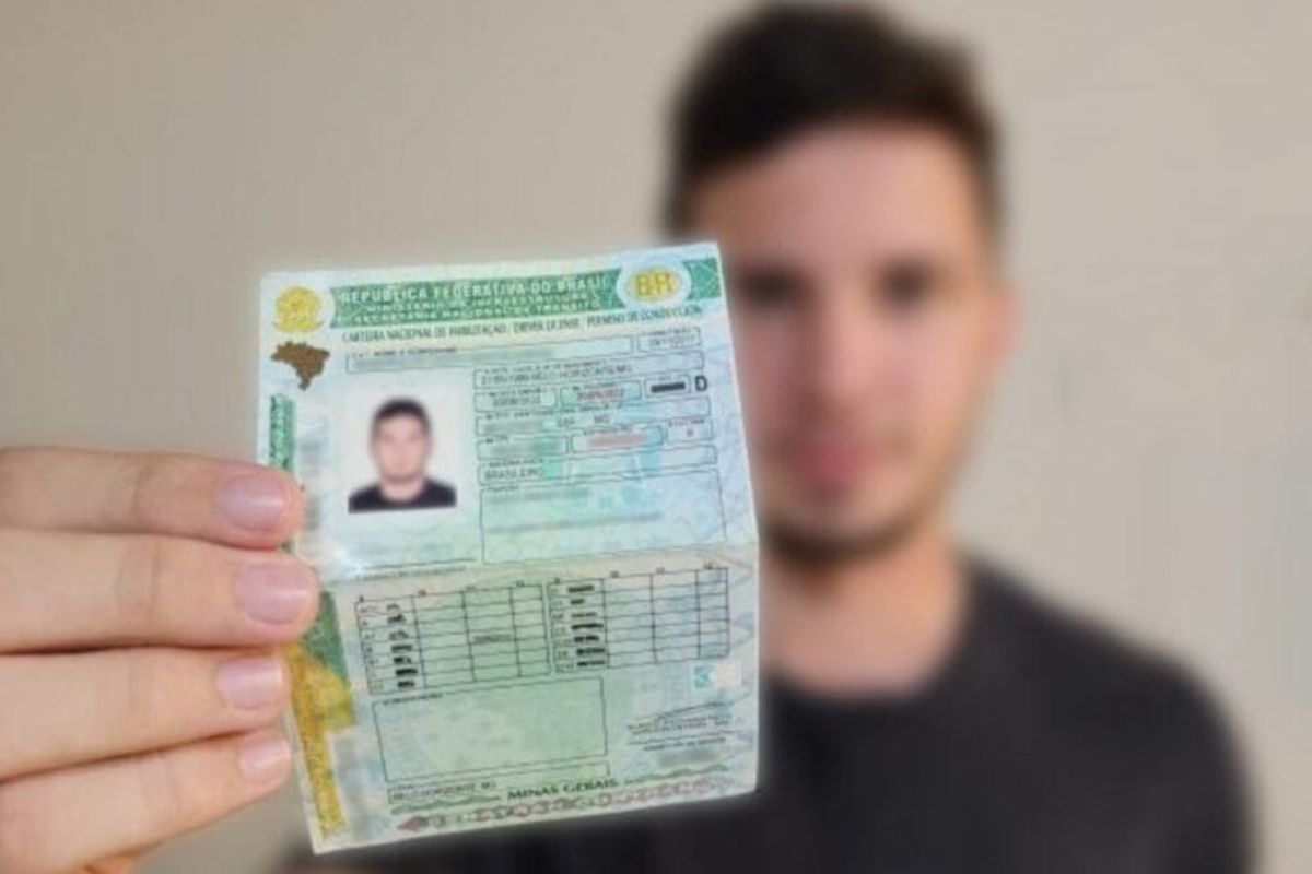 Mão segurando e exibindo uma Carteira Nacional de Habilitação (CNH) brasileira, dobrada ao meio, com o lado que contém a foto e os dados principais voltado para frente.