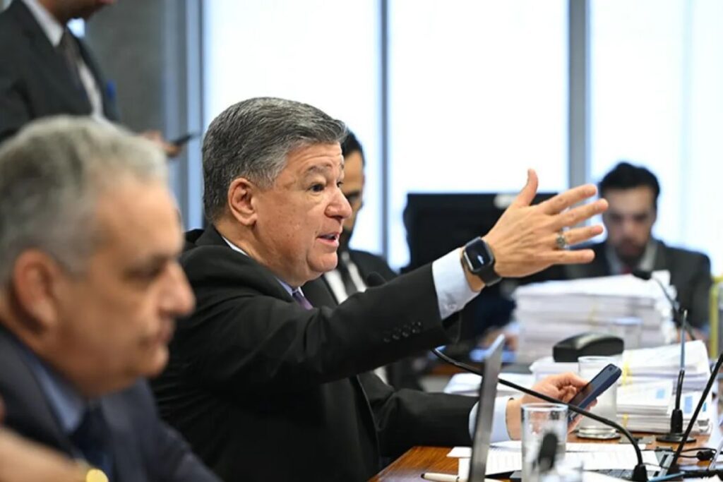 Político (provavelmente um senador ou deputado) de meia-idade, vestindo um terno escuro, fala em uma mesa de comissão parlamentar, gesticulando com a mão direita estendida.