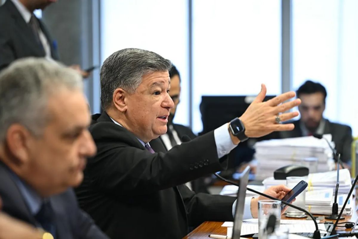 Político (provavelmente um senador ou deputado) de meia-idade, vestindo um terno escuro, fala em uma mesa de comissão parlamentar, gesticulando com a mão direita estendida.