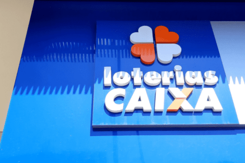 Fachada azul com o logotipo das Loterias Caixa em destaque. O símbolo apresenta um trevo de quatro folhas estilizado com corações nas cores laranja e branco, acima do nome 'Loterias CAIXA' escrito em letras brancas e laranjas em relevo.