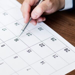 Plano geral de cima mostrando uma mão com uma caneta esferográfica de metal sobre um calendário de papel branco.