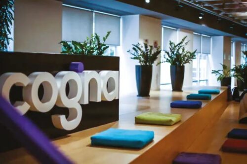 Logotipo da Cogna em letras brancas e em relevo sobre uma base escura em destaque. O ambiente é um escritório moderno e iluminado, com um longo banco de madeira adornado por almofadas coloridas em tons de azul, verde e roxo.