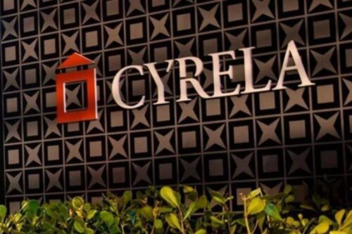 Fachada escura com o logotipo da Cyrela (uma forma estilizada de telhado e casa em um quadrado vermelho) e o nome da empresa em letras brancas grandes, iluminadas.