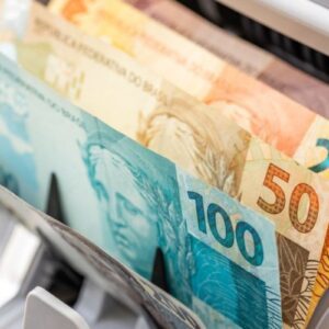 Cédulas de Real (R$), incluindo notas de R$ 100, R$ 50 e R$ 20, sendo alimentadas ou contadas por uma máquina contadora de dinheiro em um close-up.