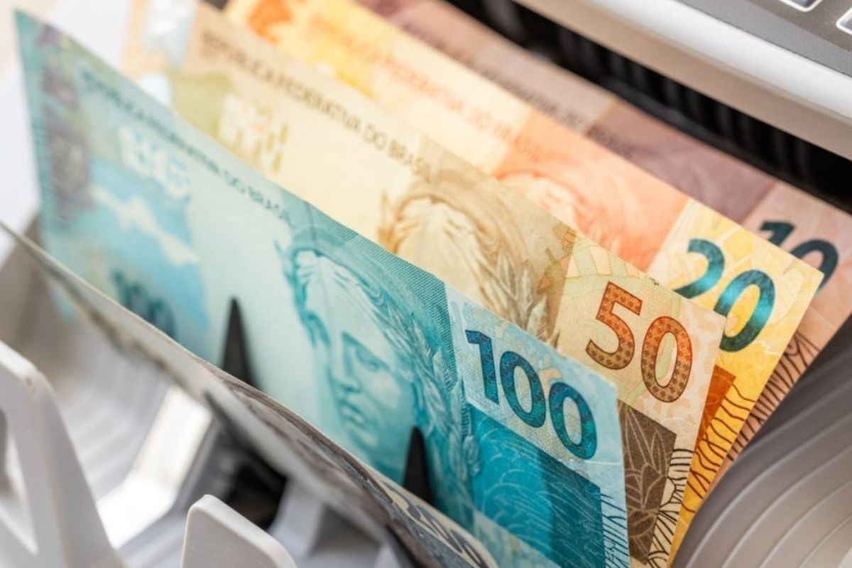 Cédulas de Real (R$), incluindo notas de R$ 100, R$ 50 e R$ 20, sendo alimentadas ou contadas por uma máquina contadora de dinheiro em um close-up.