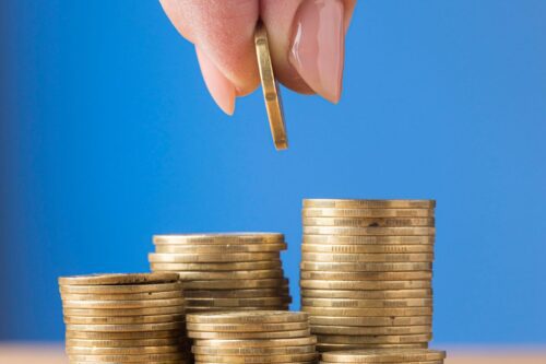 Detalhe de uma mão com unhas bem cuidadas, segurando uma moeda dourada e adicionando-a ao topo de uma das várias pilhas crescentes de moedas de ouro. O fundo é de cor azul sólida. A imagem evoca os conceitos de poupança, investimento, juros compostos ou recebimento de dividendos.