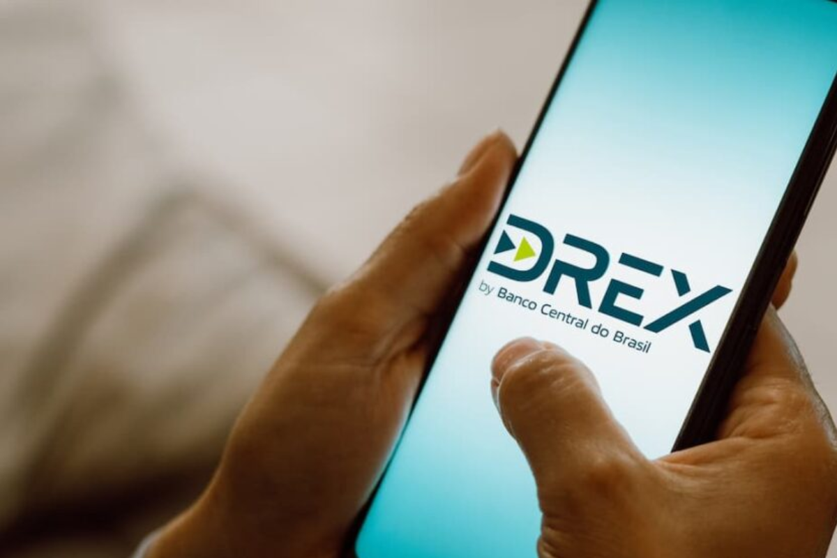lose-up de mãos segurando um smartphone que exibe na tela o logotipo oficial do Drex, a moeda digital brasileira, com a inscrição 'by Banco Central do Brasil' logo abaixo