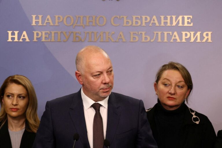 Homem careca de terno escuro, possivelmente um político, falando em um pódio com microfones. Ele está flanqueado por duas mulheres. Ao fundo, está escrito em cirílico "НАРОДНО СЪБРАНИЕ НА РЕПУБЛИКА БЪЛГАРИЯ" (Assembleia Nacional da República da Bulgária).