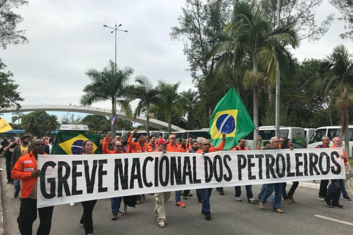 Fotografia colorida de uma manifestação de rua no Brasil. No centro, um grupo de trabalhadores veste uniformes laranjas e segura uma grande faixa branca com letras pretas onde se lê: 'GREVE NACIONAL DOS PETROLEIROS'.