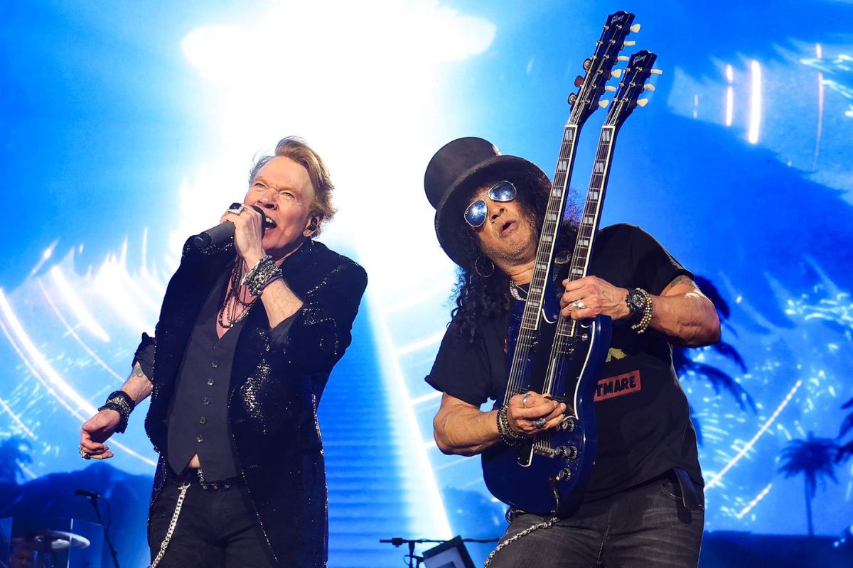 Os membros da banda Guns N' Roses, o vocalista Axl Rose e o guitarrista Slash, se apresentam em um palco iluminado por luzes azuis e brancas.