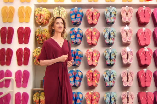 atriz Fernanda Torres em frente a uma parede de exposição com dezenas de pares de chinelos Havaianas coloridos e estampados. A imagem faz referência à Alpargatas, uma das principais empresas do portfólio da holding Itaúsa.