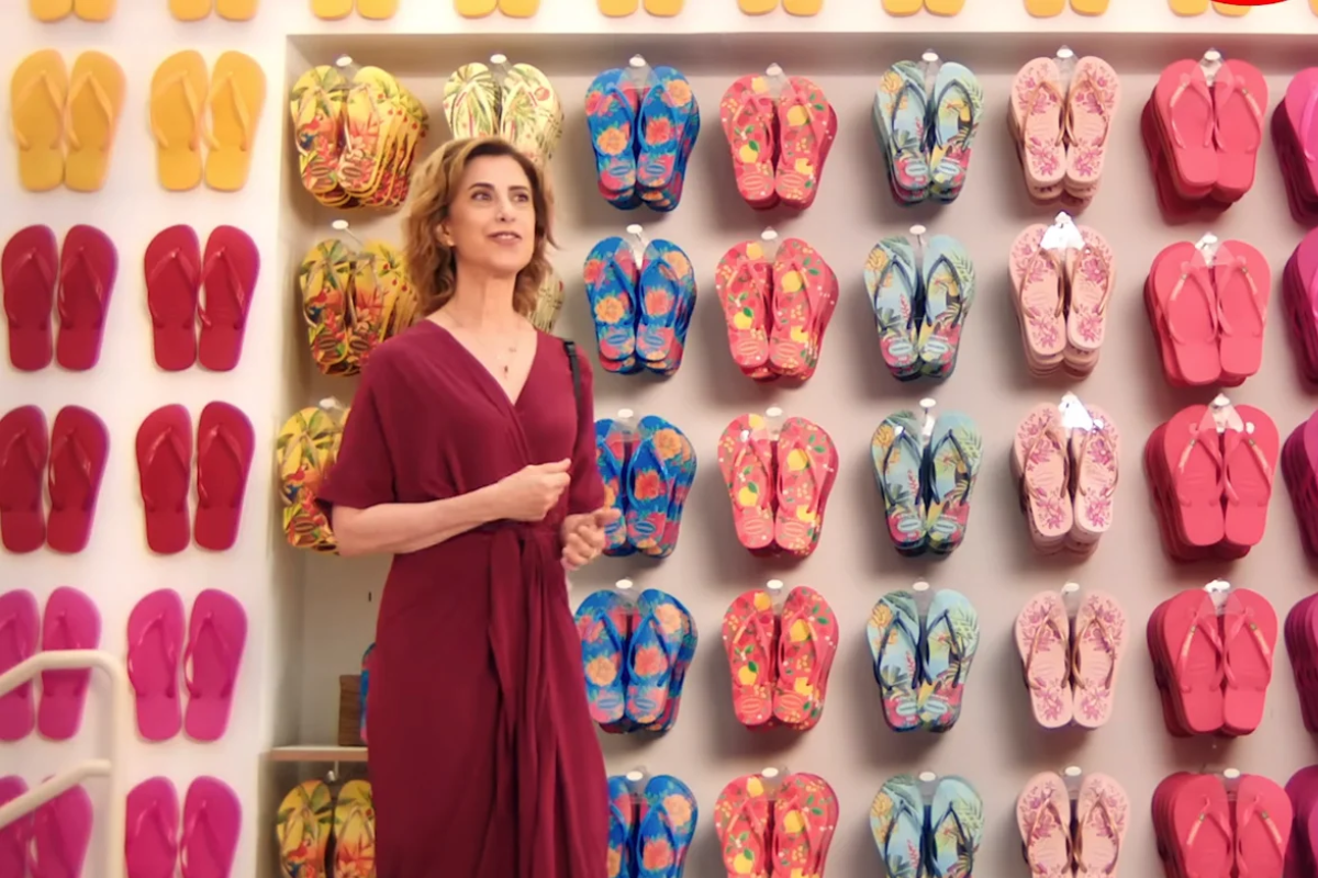 atriz Fernanda Torres em frente a uma parede de exposição com dezenas de pares de chinelos Havaianas coloridos e estampados. A imagem faz referência à Alpargatas, uma das principais empresas do portfólio da holding Itaúsa.