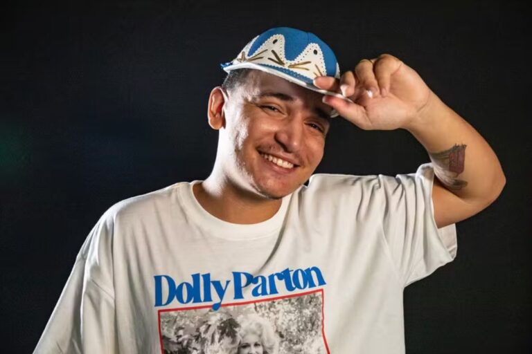 O cantor brasileiro João Gomes em foto de estúdio, sorrindo e tocando o boné azul e branco que usa.