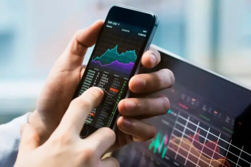 Pessoa utilizando um smartphone e um tablet simultaneamente para analisar gráficos de ações e dados financeiros.