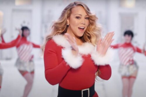 Mariah Carey sorridente e batendo palmas, vestida com um traje natalino de veludo vermelho com gola e punhos de pele branca e um cinto preto largo.