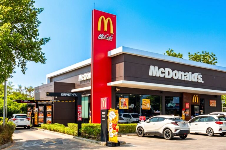 Vista externa de uma moderna loja McDonald's, com fachada em tons de marrom e branco, e uma alta torre vermelha com o logotipo amarelo e o letreiro "McCafé".