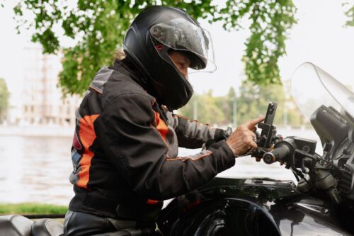 Um motociclista, usando um capacete preto integral e jaqueta de proteção preta com detalhes em laranja, está sentado em sua motocicleta.