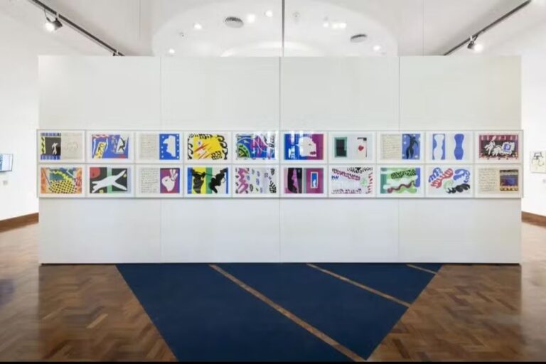 Uma parede branca de galeria de arte com duas fileiras horizontais de quadros.