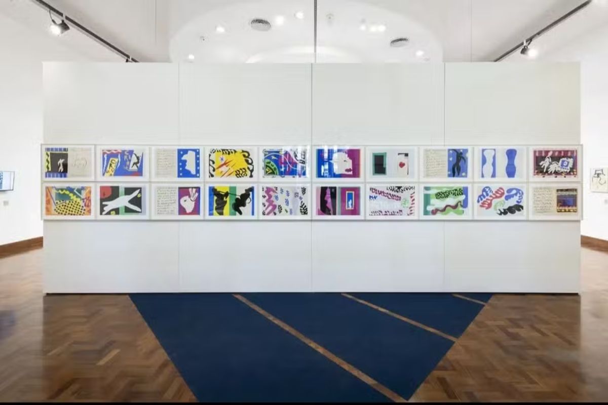 Uma parede branca de galeria de arte com duas fileiras horizontais de quadros.